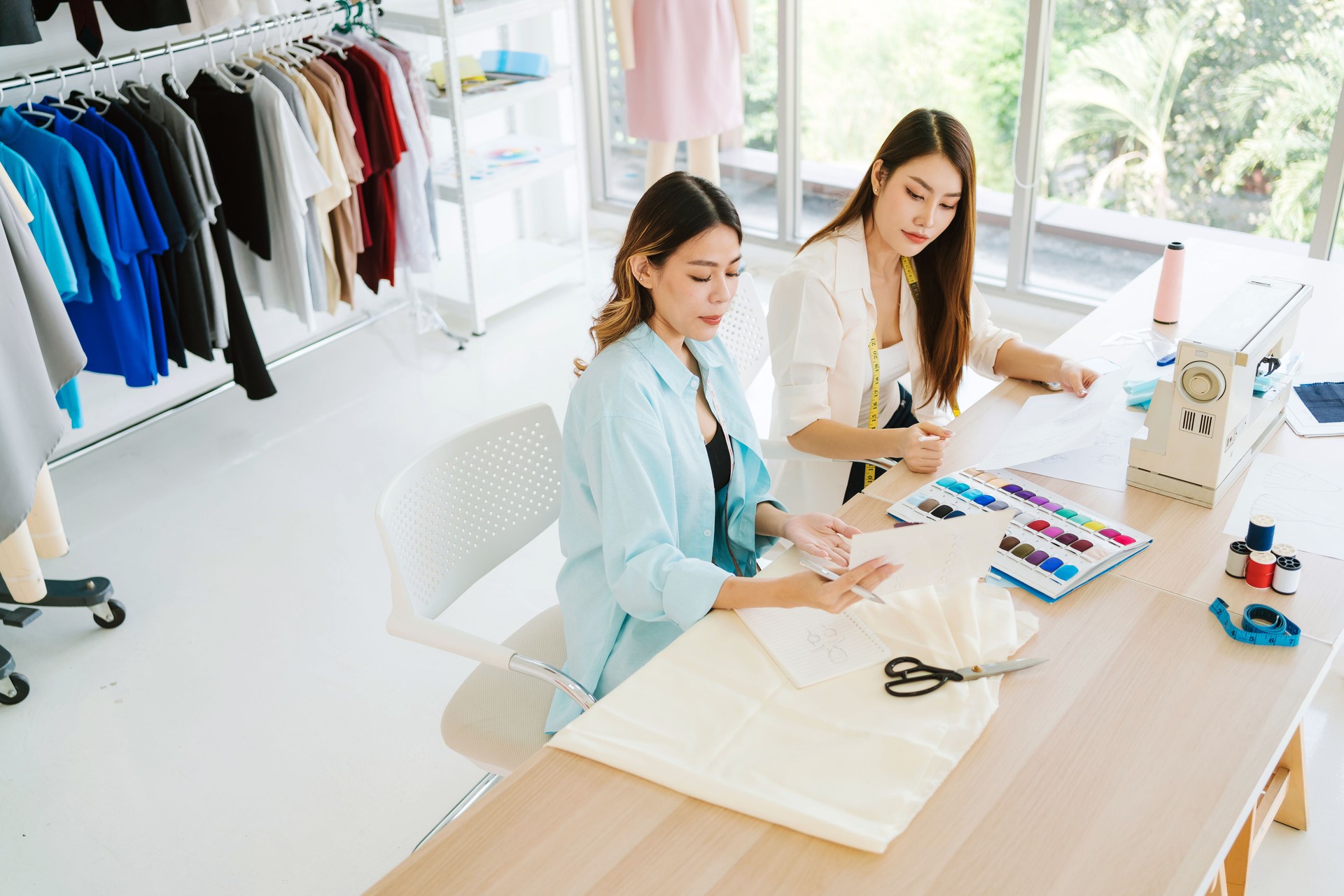 Two Asian female fashion designers or dressmakers brainstormed and discuss the design of the new collection with samples of fabrics and sketches on the laptop. Working together in the studio.