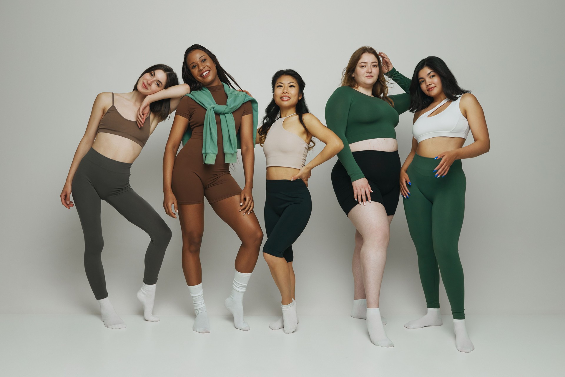 Diverse group of five women standing together in athleisure wear, showcasing different body types and ethnic against white background.